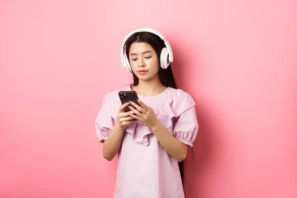 Młoda dziewczyna słuchająca muzyki w słuchawkach i rozmawiająca przez telefon komórkowy, stojąca w sukience na różowym tle — Zdjęcie stockowe
