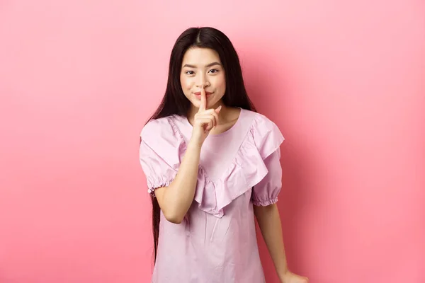 Χαριτωμένο κορίτσι της Ασίας κρύβεται μυστικό, hushing με το δάχτυλο πιέζεται στα χείλη και χαμογελά, ζητώντας να κρατήσει ήσυχο, στέκεται στο φόρεμα σε ροζ φόντο — Φωτογραφία Αρχείου
