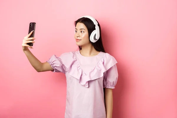 Piękna japońska dziewczyna robi selfie na smartfonie, nosi słuchawki bezprzewodowe, stoi na różowym tle — Zdjęcie stockowe