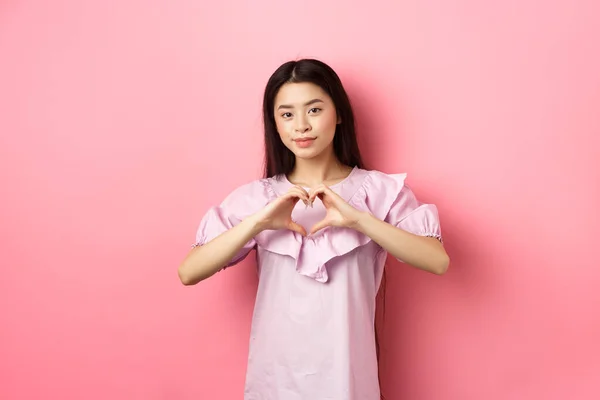 Sevgililer Günü konsepti. Güzel Japon genç kız kalp jesti ve gülümseme şirin, aşığına sevgi ve sempati göster, pembe arka planda romantik elbiseler içinde duruyor. — Stok fotoğraf