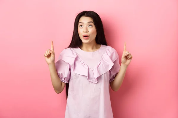 Emocionado adolescente asiático mujer apuntando los dedos hacia arriba, mirando soñador en el espacio vacío, de pie en vestido sobre fondo rosa — Foto de Stock