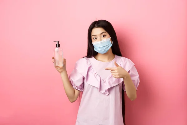 Sağlıklı insanlar ve covid-19 salgını. Tıbbi maskeli şık Japon kız el dezenfektanı şişesini gösteriyor, antiseptiğe bakıyor, pembe arka planda duruyor. — Stok fotoğraf