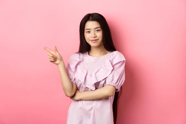 Όμορφη έφηβη Ασιάτισσα δείχνει το δάχτυλο αριστερά, χαμογελώντας με αυτοπεποίθηση, δείχνοντας διαφήμιση σε ροζ φόντο — Φωτογραφία Αρχείου