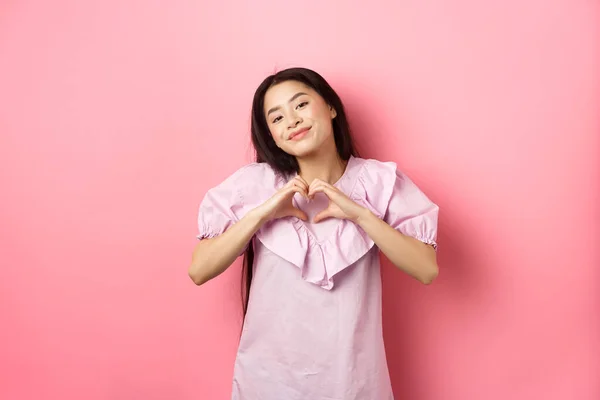 Η ιδέα του Αγίου Βαλεντίνου. Όμορφη Κορεάτισσα έφηβη με φόρεμα που δείχνει χειρονομία καρδιάς και χαμογελώντας με θαυμασμό και στοργή, ερωτεύεται, αισθάνεται ρομαντική, ροζ φόντο — Φωτογραφία Αρχείου
