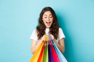 Alışveriş torbalarına heyecanla bakan, mağazalardan indirimli alışveriş yapan, mavi arka planda duran mutlu genç bir kadın.