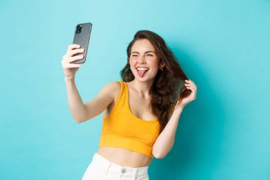Teknoloji ve yaşam tarzı konsepti. Mutlu genç bir kadın akıllı telefon uygulamasında filtreli selfie çekerken aptal suratlar yapıyor, mavi arka planda duruyor.