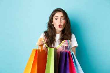 Renkli alışveriş torbaları taşıyan, alışveriş meraklısı genç bir kadın. Yan dükkandan bir şeyler alıyor, mavi arka planda dikiliyor.
