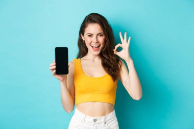 Şuna bir bak. Şımarık gülüşlü, güzel bir kadın, göz kırpan ve işaret eden boş akıllı telefon ekranlı, uygulama öneren, mavi arka planda duran