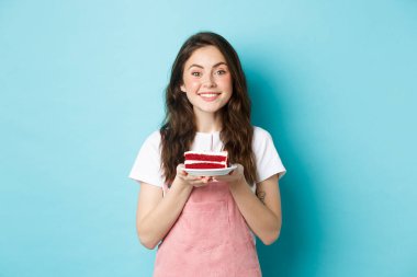 Kutlama ve tatil. Gülümseyen genç kadın doğum gününü kutluyor, elinde mumla pasta tutuyor ve kameraya rüya gibi bakıyor, mavi arka planda duruyor.