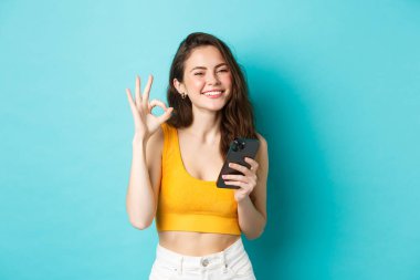 Teknoloji ve yaşam tarzı konsepti. Gülümseyen güzel bir kız, onay işareti gösteriyor, evet de, akıllı telefonu tut, mavi arka planda dur.