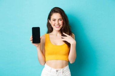 Yazlık kıyafetli beyaz kadın boş akıllı telefon ekranı ve gülümsemesi gösteriyor, bir uygulama veya internet mağazası gösteriyor, mavi arka planda duruyor.