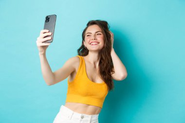 Güzel modern kız tatilde selfie çekiyor, yaz kıyafetleriyle poz veriyor ve akıllı telefon kamerasına bakıyor, mavi arka planda duruyor.