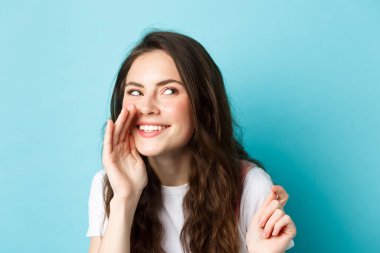 Kurnaz aptal kadın dedikoduları yayar, sessizce konuşur, dedikoduları anlatır, sağ tarafta gülümser ve kıkırdar, mavi arka planda durur.