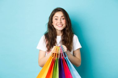 İndirimli alışveriş yapan, alışveriş torbaları tutan, mavi arka planda mutlu duran neşeli genç bir kadın.