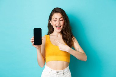 Genç mutlu güzel kız, boş akıllı telefon ekranına bakıyor ve işaret ediyor, cep telefonu ekranında reklamını gösteriyor, mavi arka planda duruyor.