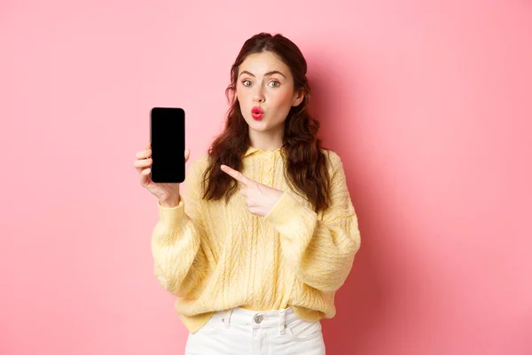 Технологии и интернет-магазины. Возбужденная девушка выглядит любопытно, указывает пальцем на пустой экран мобильного телефона, показывает приложение на смартфоне, стоя на розовом фоне — стоковое фото