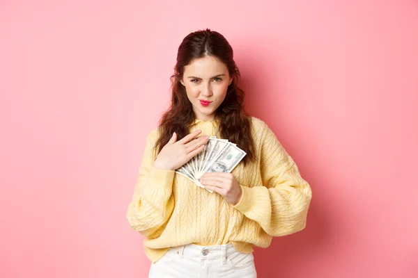 Compras. Mujer joven descarada sonriendo y mirando confiado, abrazando el dinero, sosteniendo billetes de dólar en el pecho, mirando pensativo a la cámara, de pie sobre el fondo rosa — Foto de Stock