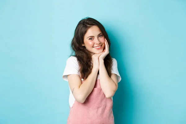 Retrato de mujer joven tonta y linda risita coqueta, sonriente y coqueta a la cámara, ruborizada, con traje de verano y maquillaje brillante, de pie sobre fondo azul — Foto de Stock