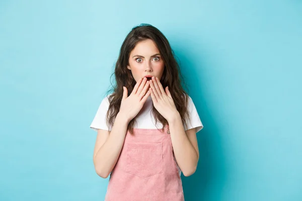 Portrait de commérage choqué fille haletant, couvrant la bouche ouverte avec les mains et le regard surpris à la caméra, entendre la rumeur, regardant avec incrédulité, debout sur fond bleu — Photo