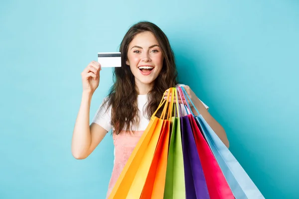 Mulher bonita feliz mostrando cartão de crédito de plástico e sacos de compras com mercadorias, comprando com descontos, de pé sobre fundo azul — Fotografia de Stock