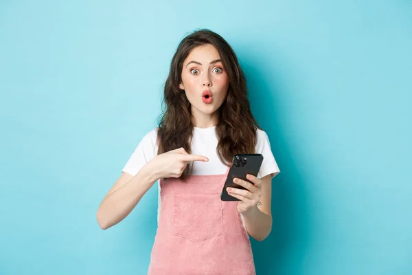 Porträtt av upphetsad kvinna pekar finger åt smartphone, berättar om online promo erbjudande, visar reklam på telefon, står mot blå bakgrund — Stockfoto