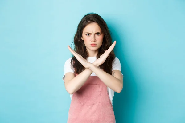 Image de jeune femme déçue fronçant les sourcils, faisant signe de croix et boudant, montrant geste d'arrêt, dire non, interdire la mauvaise action, debout sur fond bleu — Photo