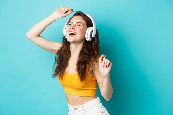 Hermosa chica de verano bailando y riendo en auriculares, escuchando música con sonrisa despreocupada, de pie sobre fondo azul — Foto de Stock