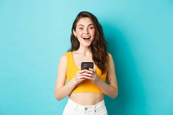 Ung upphetsad kvinna får goda nyheter på telefon, hålla smartphone, titta förvånad över kameran med glada leende, står mot blå bakgrund — Stockfoto