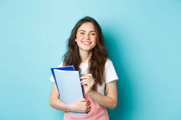 Boldog fiatal lány diák kezében notebookok tanfolyamok és mosolygó kamera, álló tavaszi ruhák ellen kék háttér — Stock Fotó