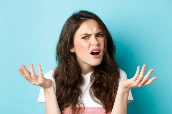 Immagine di una giovane donna confusa e infastidita che scrollava le spalle, chiedendosi cosa fosse, fissando interrogata e dispiaciuta la macchina fotografica, in piedi su uno sfondo blu — Foto Stock