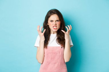 Angry and pissed-off brunette woman shaking hands and frowning outraged, look with hatred, standing furious and aggressive against blue background
