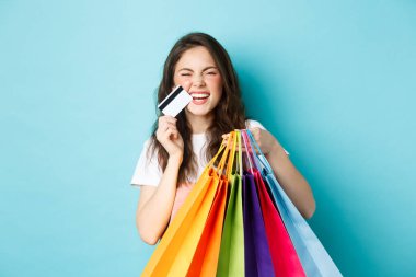 Mutlu genç bir kadın gülüyor, alışveriş torbaları ve plastik kredi kartları gösteriyor, indirimli mağazalardan alışveriş yapıyor, mavi arka plana karşı neşeli duruyor.