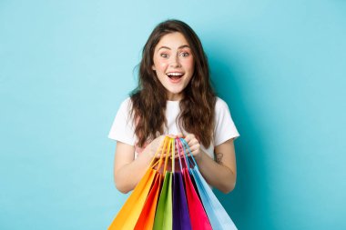 Heyecanlı kız alışverişçinin portresi, alışveriş torbaları, mağazalardan alışveriş yapmak, şaşırmış bir şekilde gülümsemek, indirimlerden hoşlanmak, mavi arka planda dikilmek.
