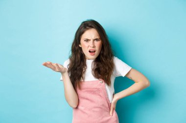 Image of confused and annoyed young woman raising hand and pointing at something terrible, staring puzzled at camera with pissed-off face, standing over blue background