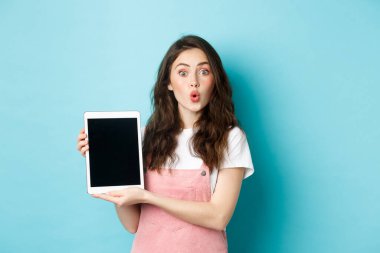Şaşırmış çekici bir kadın dijital tablet ekranı gösteriyor ve kameraya hayret içinde bakıyor, tanıtım teklifini kontrol ediyor, mavi arka planda eğleniyor.