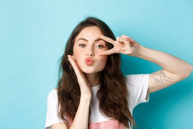 Mükemmel cilde sahip güzel genç bir bayana yakın durun. Göze yakın barış işareti ve öpüşen bir yüz gösterin. Mavi arka planda durun.