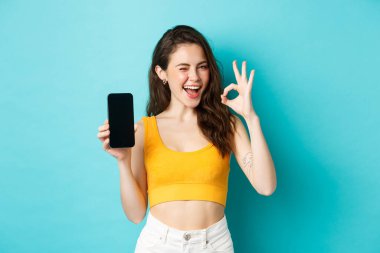 Yaz kıyafetleri içinde çekici bir modern kadın, güven verici bir şekilde göz kırpıyor, boş akıllı telefon ekranıyla işaret dilini gösteriyor, logonuzu ekranda gösteriyor, mavi arka plan.