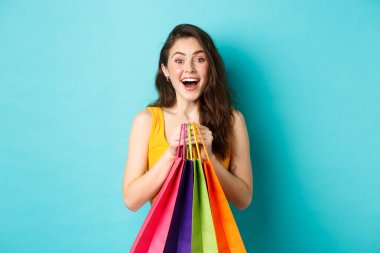 Şaşırmış genç bir kadının portresi kameraya hayret içinde bakıyor, elinde alışveriş torbaları, mağazada indirimler görüyor, mavi arka planda duruyor.