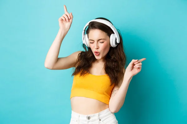 Elegante mujer moderna en auriculares, escuchando música y bailando, disfrutando de canciones favoritas en auriculares, de pie sobre fondo azul — Foto de Stock