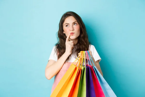 Προσεκτική γυναίκα κρατώντας τσάντες για ψώνια, αγγίζοντας το πρόσωπο και να κοιτάξουμε μακριά, ενώ σκέφτεται, κάνοντας επιλογή στο κατάστημα, στέκεται πάνω από το μπλε φόντο — Φωτογραφία Αρχείου