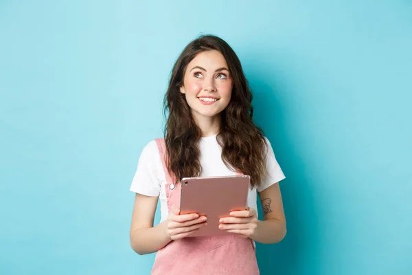 Menina sonhadora bonita sorrindo, segurando tablet nas mãos e olhando para o logotipo do canto superior esquerdo com rosto pensativo, de pé sobre fundo azul — Fotografia de Stock