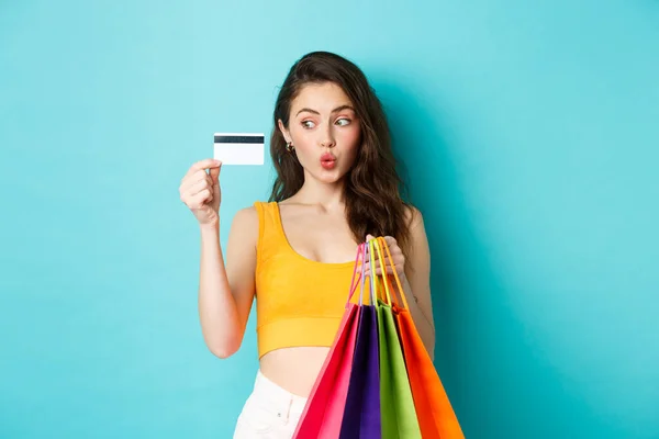 快乐的女性购物狂展示她的塑料信用卡，拿着购物袋，穿着夏装，站在蓝色背景下的照片 — 图库照片