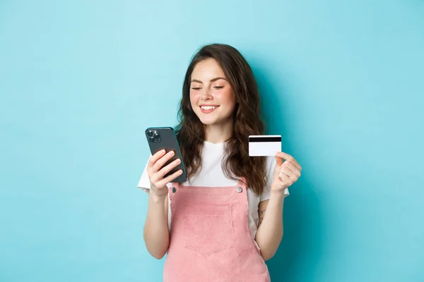 Joven chica moderna que paga en línea con tarjeta de crédito, tipo de información en el teléfono inteligente, mirando a la pantalla con la cara complacida, compras en la aplicación, de pie sobre fondo azul — Foto de Stock