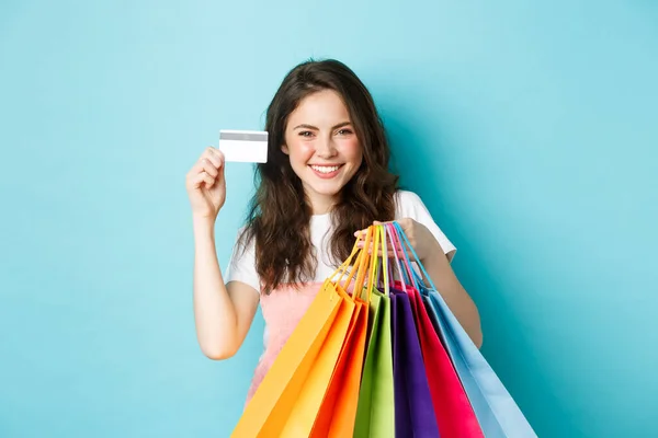 Mladá spokojená žena se usmívá, ukazuje plastové kreditní karty a drží nákupní tašky, nákup s bezkontaktní platbou, stojící nad modrým pozadím — Stock fotografie