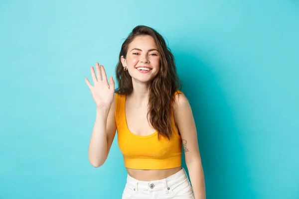 Summer holidays and emotions concept. Beautiful friendly woman with perfect body, waving hand and saying hi, greeting you, standing over blue background