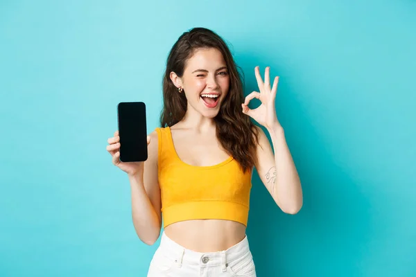 Atrakcyjna nowoczesna kobieta w letnich ciuchach, mrugająca pewnie, pokazująca w porządku znak z pustym ekranem smartfona, pokazująca logo na wyświetlaczu, niebieskie tło — Zdjęcie stockowe
