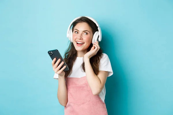 Obraz młodej kobiety zakładającej słuchawki i wybierającej muzykę na smartfona, trzymającej telefon, słuchającej piosenki, stojącej nad niebieskim tłem — Zdjęcie stockowe