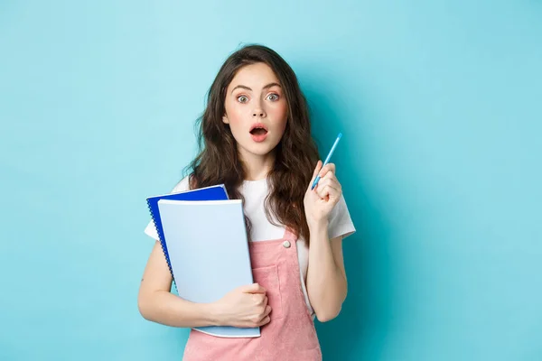 Got an idea. Excited young woman student, holding notebooks and pen, raising hand while having a plan, think-up good solution, standing over blue background