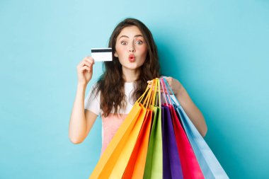 Plastik kredi kartı ve alışveriş torbaları gösteren, öpüşmek için dudaklarını büzen, mavi arka planda mutlu duran çekici kız.