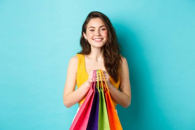 Alışverişe giden neşeli bir kızın resmi. Elinde alışveriş poşetleri ve kameraya gülümsüyor. Mavi arka planda duruyor.